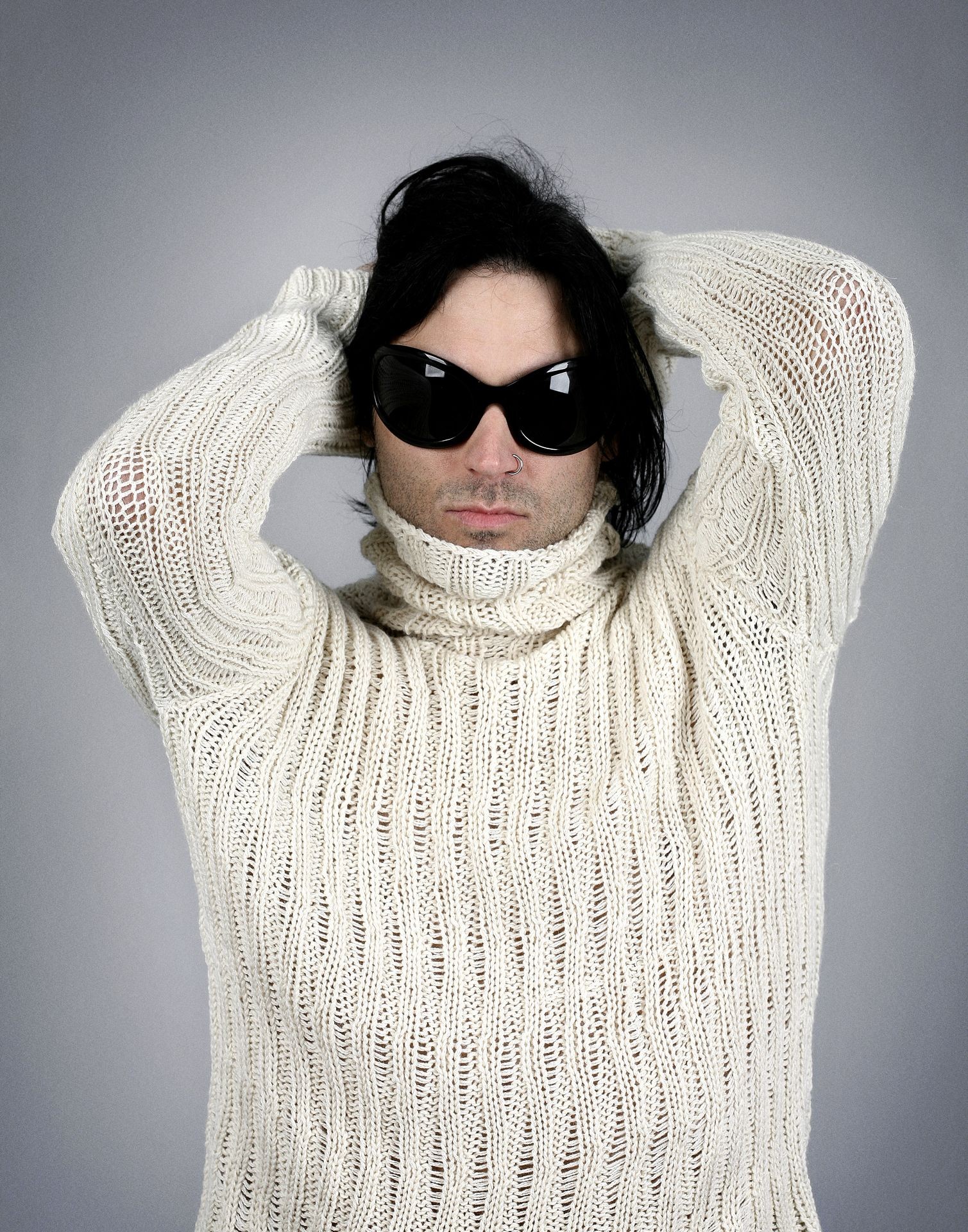 Individual wearing a light-colored, knitted sweater with arms raised and hands resting behind their head.