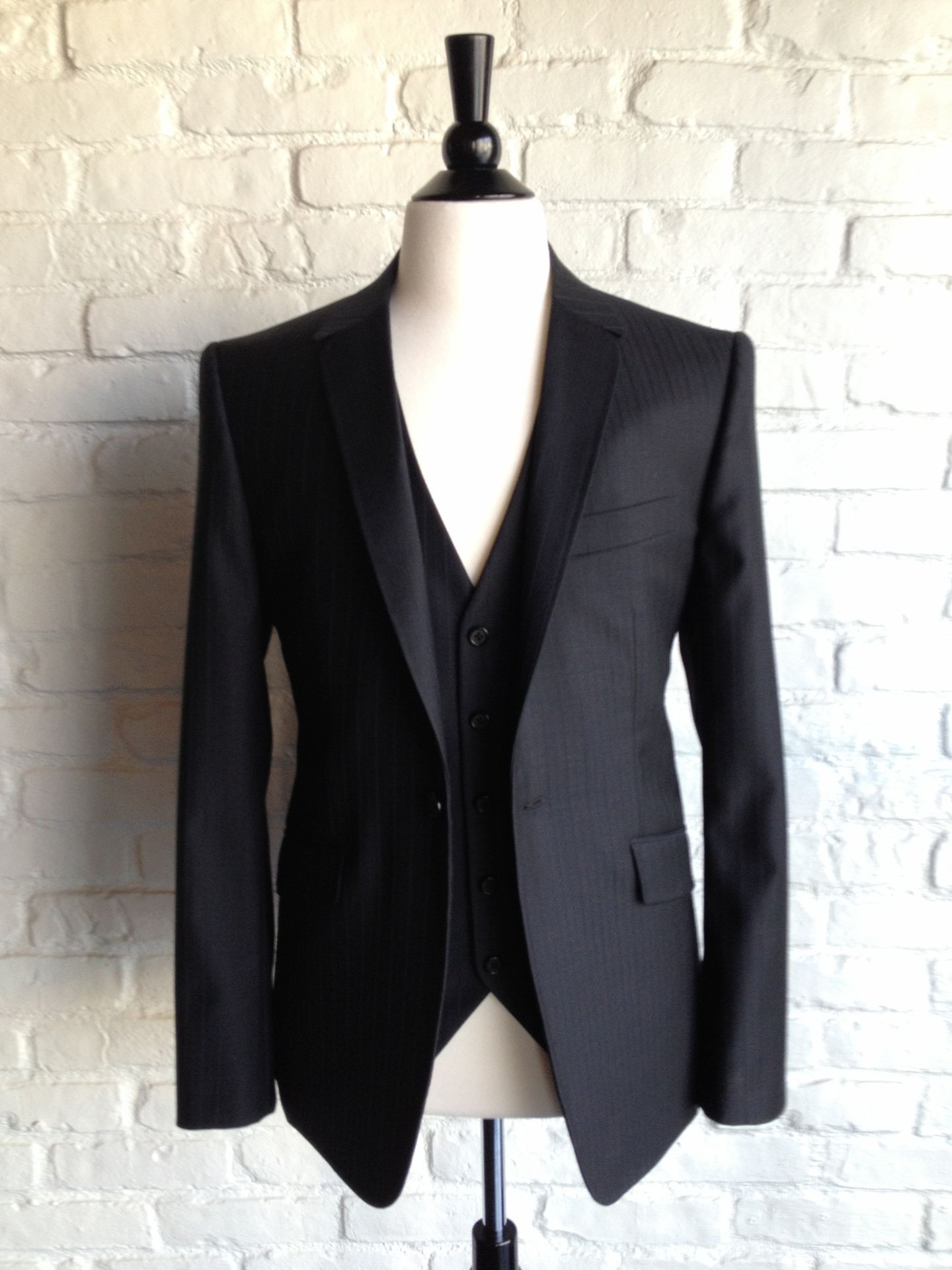 Black three-piece suit displayed on a mannequin in front of a white brick wall.